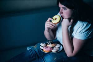 Unhappy woman sitting in dark, watching tv, eating a plate of donuts.